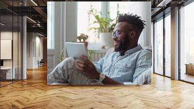 Relaxed smiling african american man holding digital tablet computer using apps sitting on couch at home. Black guy remote learning, social distance working, ordering buying online or reading e book. Wall mural