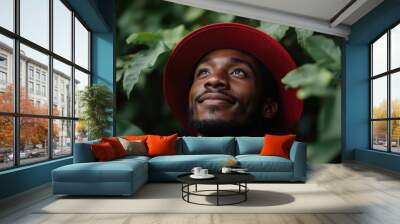 A bearded man wearing a red hat, looking up amidst lush green leaves in a natural setting, radiating curiosity and contemplation in a tranquil environment. Wall mural