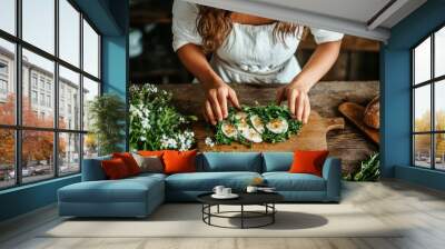 A person diligently arranges food on a wooden chopping board inside, surrounded by fresh greenery, ingredients, and rustic decor, highlighting home-style cooking’s art. Wall mural