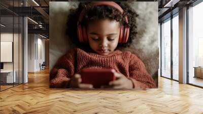 A young child is wearing red headphones and is deeply engrossed in using a smartphone while lounging on a soft chair, highlighting the engagement and immersion of digital age interactions. Wall mural