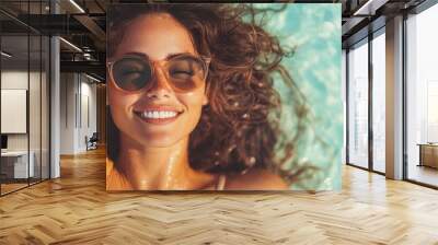 A young woman with curly hair, wearing sunglasses, smiles peacefully while reclining in clear blue water, capturing a moment of joy and relaxation under the sun. Wall mural