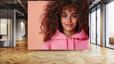 A young woman with curly hair wearing a vibrant pink hoodie smiles softly, with an orange background adding warmth and positive vibes to the image. Wall mural