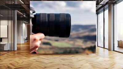 A close-up of a woman s hand holding a camera, with the lens focused on a landscape Wall mural