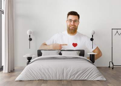 Happy smiling bearded male model wears casual white t shirt, round spectacles, indicates at red heart sewed on clothes, isolated over white studio background. Cheerful man advertizes new outfit Wall mural