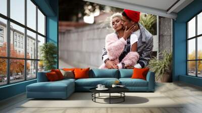 Loving couple embrace each other while have walk outdoor, kiss. Dark skinned stylish male in trendy red hat hugs his girlfriend with specific appearance. People, ethicity, relationship concept Wall mural