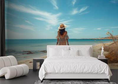 a young pretty woman in a hat and a light dress is walking on the sandy beach. Wall mural