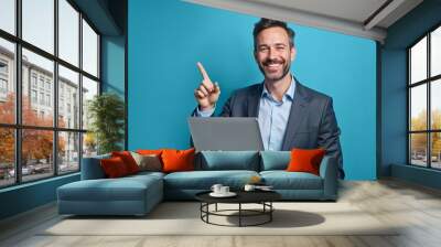 A cheerful businessman holding a laptop and pointing upwards, symbolizing success and innovation. Ideal for corporate presentations, motivational content, or online courses Wall mural