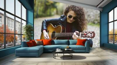 Beautiful young woman playing guitar sitting on the forest. Girl wearing black jacket and sunglasses, fashion lifestyle. Wall mural