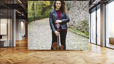 Portrait of beautiful smiling woman posing with a guitar on forest. Girl wearing black jacket, fashion lifestyle. Wall mural