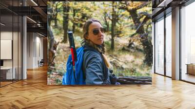 A woman wearing sunglasses and a blue backpack is sitting on a log in a forest. She is smiling and she is enjoying her time outdoors Wall mural