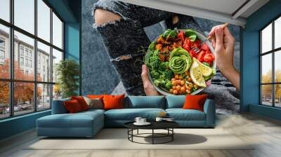 Woman in jeans holding Buddha bowl with salad, baked sweet potatoes, chickpeas, broccoli, greens, avocado, sprouts in hands. Healthy vegan food, clean eating, dieting, top view Wall mural