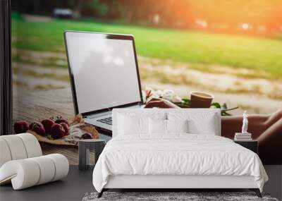 Young woman using and typing laptop computer at rough wooden table with coffee cup, strawberries, bouquet of peonies flowers, smartphone. Freelancer working in outdoor park. Toning Wall mural