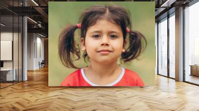 A little girl with a confident look looks into the frame on a green natural background, a brunette child with big brown eyes Wall mural