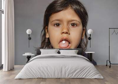 Close-up portrait of a little funny charming funny caucasian girl with a pill on her tongue on a gray background, taking medicine, taking vitamins, tongue, eating, being treated, eating, health care Wall mural