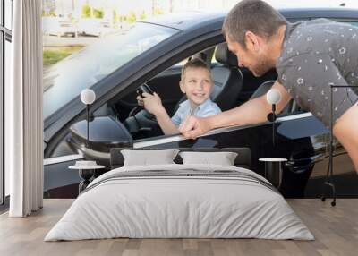 Cute boy is sitting at the wheel in the car, dad is looking out the car window Wall mural