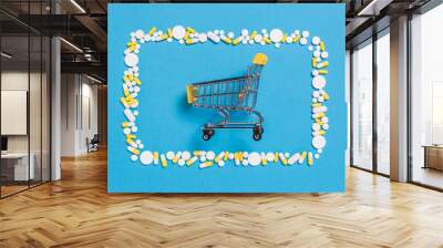 Yellow shopping cart in frame of white pills and capsules on blue pharmaceutical background. Buying medicine Wall mural