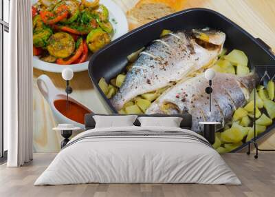 Two baked fish and potato in pan at wooden table with sauce and vegetables Wall mural