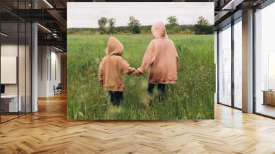 Two children walk in a field of winter crops in sweatshirts with hoods and boots, freedom in nature, back view Wall mural