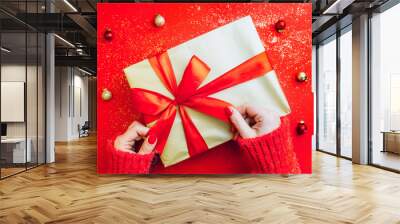 Female hands holding present with red bow on red sparkling background. Festive backdrop for Christmas or New Year. Flat lay. Top view Wall mural