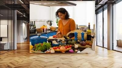 Young brunette woman cutting a fresh tomato on a cutting board in the kitchen at home Wall mural