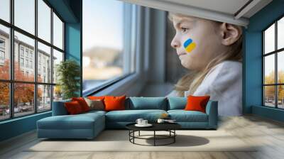 a crying child, looking out the window, with painted on her cheek the yellow-blue colours of the Ukrainian flag, a request for help. Children hope for peace. Wall mural