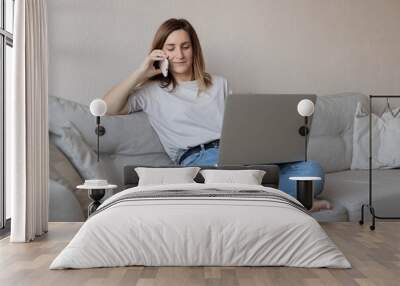 Portrait of female freelancer working at home with laptop and mobile phone. Happy woman surfing and browsing net and talking on smartphone, sitting on couch, relaxing in living room. Wall mural