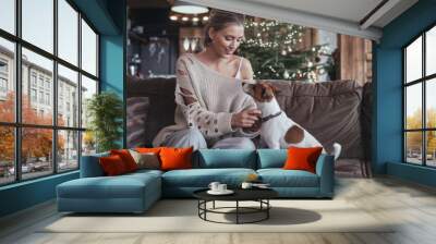 Woman sitting on a couch with her dog in a cozy home environment. Christmas tree with lights in the background. Holiday season concept. Wall mural