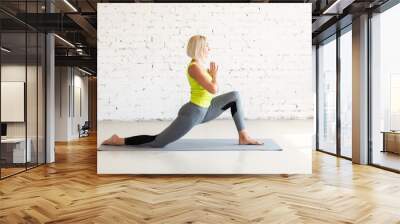 Hip joint mobility. Fit adult caucasian woman practice stretching in deep lunge on a mat in loft white studio indoor, selective focus. Wall mural