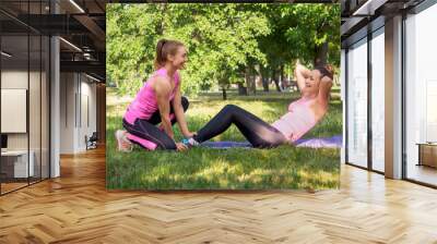 Workout with trainer. Smiling caucasian woman does crunches with the help of a coach in public park in summer, selective focus Wall mural