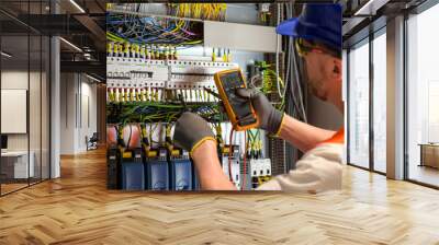 electrician in blue helmet, yellow glasses and orange vest measures electric current with digital multimeter in distribution box-blurred background Wall mural