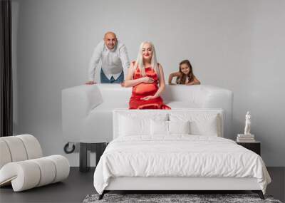 Beautiful pregnant mother in a red dress and father with a cute and emoticon girl on a white sofa isolated on a white background. Family, baby birth concept Wall mural