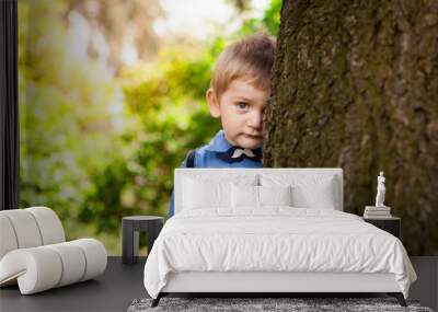 Cute little boy leaning against big tree and shy in summer, child portrait outdoors Wall mural