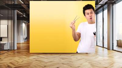 Young handsome trendy asian man eating yummy hot and spicy instant noodle using chopsticks and  bowl  isolated on yellow background in studio With copy space. Wall mural