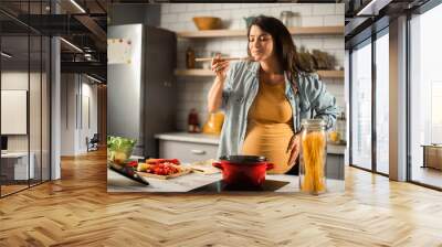 Beautiful pregnant woman preparing delicious food. Smiling woman cooking pasta at home. Wall mural