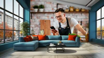 Young man in kitchen. Chef using phone while making food.  Wall mural