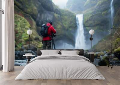 A photographer sets up camera on a tripod back turned as they capture the beauty of a pristine waterfall nestled deep . . Wall mural