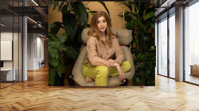 Beautiful young woman sits on the cozy chair on the terrace with tropical green plants. Portrait of model girl inside with tropical plants. Hobby and Gardening. Wellness and spa. Selective focus. Wall mural