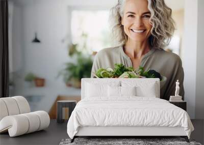 Aged woman smiling happily and holding a healthy vegetable salad bowl on blurred kitchen background, with copy space. Wall mural