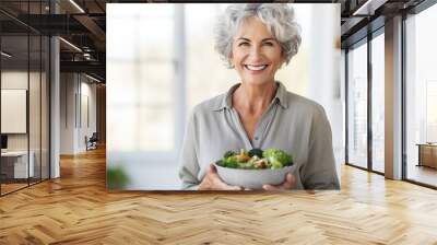 Aged woman smiling happily and holding a healthy vegetable salad bowl on blurred kitchen background, with copy space. Wall mural