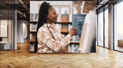 Religion book, education and black woman in a library for research, studying God and knowledge on the bible at college. Learning, smile and African student with decision of books on a scholarship Wall mural