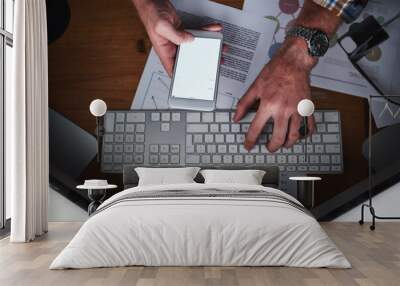 Hands on a computer keyboard and typing on a phone in an office with paper work from above. Closeup of a business man browsing the internet, finishing a report or sending a communication email Wall mural