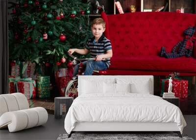 Smiling little boy riding bicycle in the room with christmas tree , new year presents , red coach, book shelf in photo studio Wall mural