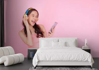 Charming asian woman wear headphones holding mobile phone listen music song mood enjoy on pink background. Joyful young girl with color headset laughing list melody radio on smartphone over isolated. Wall mural