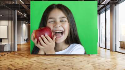 Happy child girl eat apple isolated on green background. Kid with fruits. Wall mural