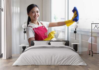 Young female cleaner is spraying cleaning solution on the glass and is wiping the window glass at home with a towel. Wall mural