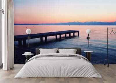 A peaceful beach with a small wooden pier extending into the water. The calm, reflective surface of the ocean and the soft light of sunset create a serene and contemplative scene Wall mural