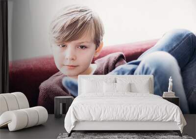 a portrait of a cute boy kid sitting on a sofa against the light window, blurred foreground with legs crossed,  soft focus image Wall mural