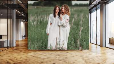 two girlfriends in the field have fun; girls in national clothes; two girls are wearing long white shirts; brunette and blonde hugging Wall mural