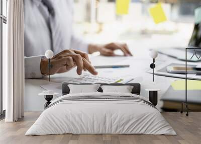 A businessman is using a white calculator to calculate financial numbers, he is sitting in his private office, the businessman examines the financial data from the corporate finance chart. Wall mural