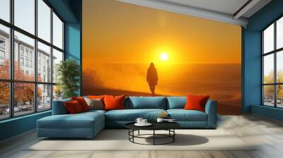 Silhouette of a person walking along the shoreline at sunset, with the ocean and sky in soft golden hues Wall mural